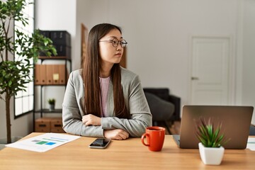 Young chinese business worker wearing business style sitting on desk at office looking to side, relax profile pose with natural face with confident smile.