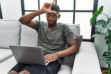 Young african american man using laptop at home sitting on the sofa making fun of people with...