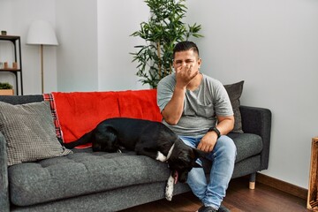 Young latin man and dog sitting on the sofa at home smelling something stinky and disgusting, intolerable smell, holding breath with fingers on nose. bad smell - obrazy, fototapety, plakaty