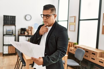 Young latin businessman with serious expression reading paperwork standing at the office.
