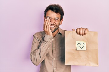 Handsome man with beard holding delivery paper bag with heart reminder laughing and embarrassed giggle covering mouth with hands, gossip and scandal concept