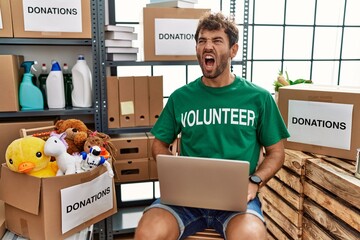 Young handsome man wearing volunteer t shirt using laptop angry and mad screaming frustrated and...