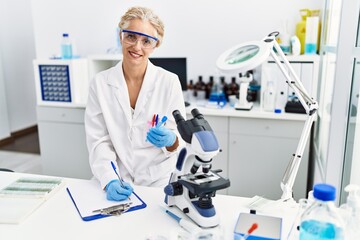 Middle age blonde woman wearing scientist uniform holding test tube writing on clipboard at laboratory