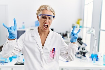 Middle age blonde woman working at scientist laboratory shouting with crazy expression doing rock symbol with hands up. music star. heavy concept.