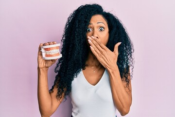 Middle age african american woman holding denture covering mouth with hand, shocked and afraid for mistake. surprised expression