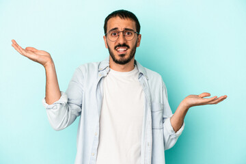 Young caucasian man isolated on blue background confused and doubtful shrugging shoulders to hold a copy space.