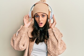 Young hispanic woman listening to music using headphones in shock face, looking skeptical and sarcastic, surprised with open mouth