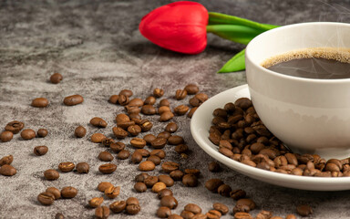 A close-up of hot coffee in a white cup is placed on a cement floor table, lots of roasted coffee beans are in the coffee cup saucer, and around, smoke and aroma waft from the cup. Blurred background