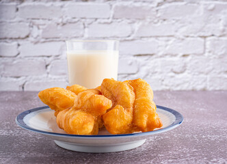 Close-up of deep-fried dough sticks or Chinese doughnut sticks on white plate and a glass of soybean milk on gray stone background with white brick wall. Space for text