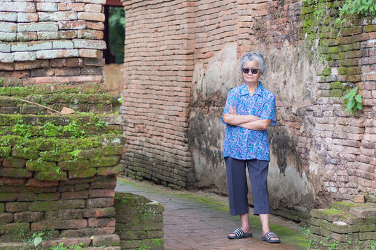Portrait of an elderly Asian woman wearing sunglasses, arms crossed and looking at the camera with a smile while standing old brick wall background. Aged people and travel concept