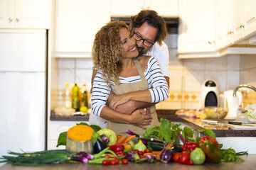 Loving man embracing his wife from behind in domestic kitchen. Man hugging wife in kitchen with variety of fresh raw vegetables on kitchen countertop at home. Romantic couple spending time in kitchen