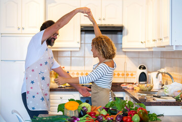 Joyful couple in apron holding hands and dancing together in domestic kitchen at home. Man and woman having fun dancing wearing apron in kitchen. Dancing caucasian couple with raw vegetable on kitchen