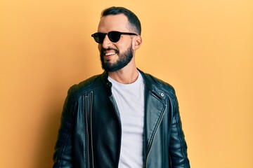 Fototapeta premium Young man with beard wearing black leather jacket and sunglasses looking away to side with smile on face, natural expression. laughing confident.