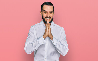 Young man with beard wearing business shirt praying with hands together asking for forgiveness smiling confident.