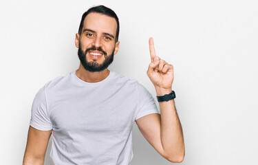 Young man with beard wearing casual white t shirt showing and pointing up with finger number one while smiling confident and happy.