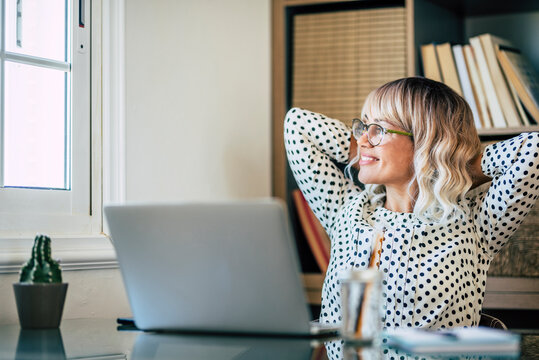 Businesswoman relaxing on chair with hands behind head while working on laptop at office. Caucasian young woman looking out through window. Woman taking a break from work and admiring view from office