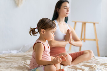 Young mom practices yoga on couch with her little daughter. Lotus position meditation. Padmasana...