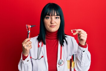 Young hispanic doctor woman holding deafness headset and otoscope relaxed with serious expression...