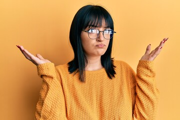Young hispanic woman wearing casual clothes and glasses clueless and confused with open arms, no...