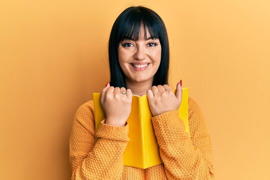 Young hispanic woman holding book smiling with a happy and cool smile on face. showing teeth.