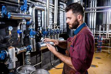 Young male brewer in leather apron supervising the process of beer fermentation at modern brewery factory