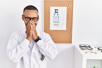 African american optician man standing by eyesight test laughing and embarrassed giggle covering...