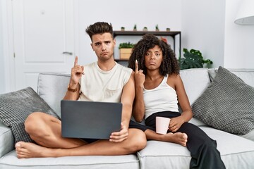 Young interracial couple using laptop at home sitting on the sofa pointing up looking sad and...