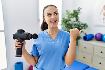 Young physiotherapist woman holding therapy massage gun at wellness center pointing thumb up to the side smiling happy with open mouth