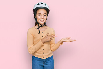 Beautiful brunette little girl wearing bike helmet amazed and smiling to the camera while presenting with hand and pointing with finger.