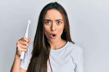 Young hispanic girl holding electric toothbrush scared and amazed with open mouth for surprise, disbelief face