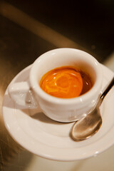 Close up. The small white ceramic mug (cup) with Italian espresso (coffee) on a table with a saucer and a metal spoon in a local Italian bar (coffee shop) in Milan, Lombardy, Italy. Beverage.