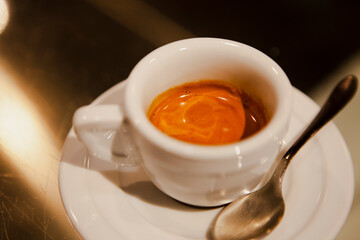 Close up. The small white ceramic mug (cup) with Italian espresso (coffee) on a table with a saucer and a metal spoon in a local Italian bar (coffee shop) in Milan, Lombardy, Italy. Beverage.