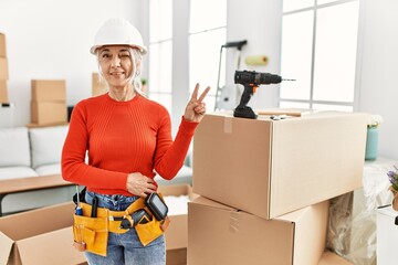 Middle age grey-haired woman wearing hardhat standing at new home smiling with happy face winking at the camera doing victory sign. number two.