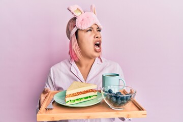 Hispanic woman with pink hair wearing pajama holding healthy breakfast angry and mad screaming frustrated and furious, shouting with anger looking up.