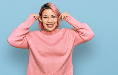 Hispanic woman with pink hair wearing casual winter sweater smiling pulling ears with fingers,...