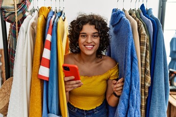 Young hispanic woman searching clothes on clothing rack using smartphone with a happy and cool smile on face. lucky person.