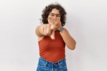 Young hispanic woman wearing glasses standing over isolated background laughing at you, pointing finger to the camera with hand over mouth, shame expression