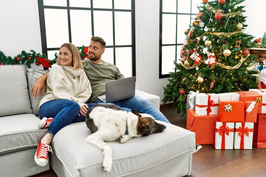 Young couple using laptop sitting by christmas tree looking away to side with smile on face, natural expression. laughing confident.