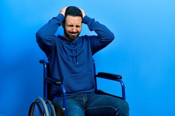 Handsome hispanic man with beard sitting on wheelchair suffering from headache desperate and stressed because pain and migraine. hands on head.