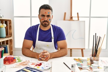 Young hispanic man with beard at art studio with painted face skeptic and nervous, frowning upset...