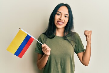 Young hispanic girl holding colombia flag pointing thumb up to the side smiling happy with open mouth