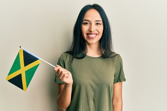 Young hispanic girl holding jamaica flag looking positive and happy standing and smiling with a confident smile showing teeth