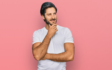 Young hispanic man wearing casual white t shirt with hand on chin thinking about question, pensive expression. smiling with thoughtful face. doubt concept.