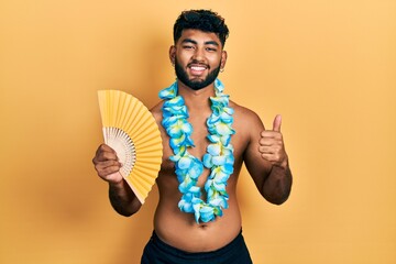 Arab man with beard wearing hawaiian lei holding handfan smiling happy and positive, thumb up doing...