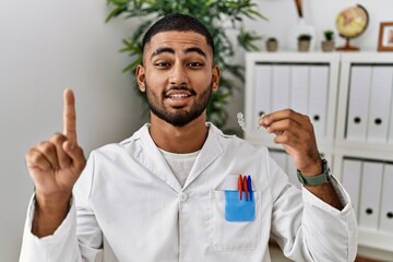 Young indian man working at dentist clinic holding invisible aligner smiling with an idea or question pointing finger with happy face, number one