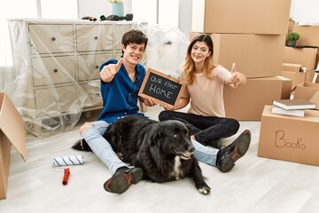 Young caucasian couple with dog holding our first home blackboard at new house approving doing...