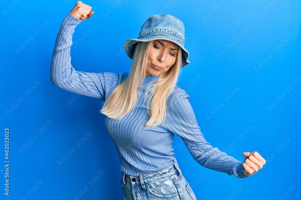 Sticker young blonde woman wearing casual denim hat showing arms muscles smiling proud. fitness concept.