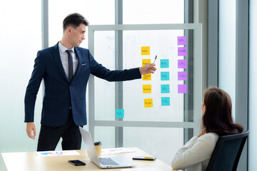 Handsome caucasian businessman are presentations of information business plans and strategy on a glass board with sticky notes to business woman are Sitting at desk with a laptop and a cup of coffee.