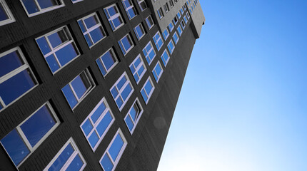 Black and white residential high-rise building with many glass windows standing against background of blue sky and sun. Urban real estate stock photo