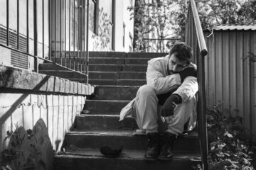 Fototapeta premium Young hungry sick and depressed homeless man sitting on the stairs of the building in the city street to rest after searching for food and to be alone out of the crowd, social documentary concept